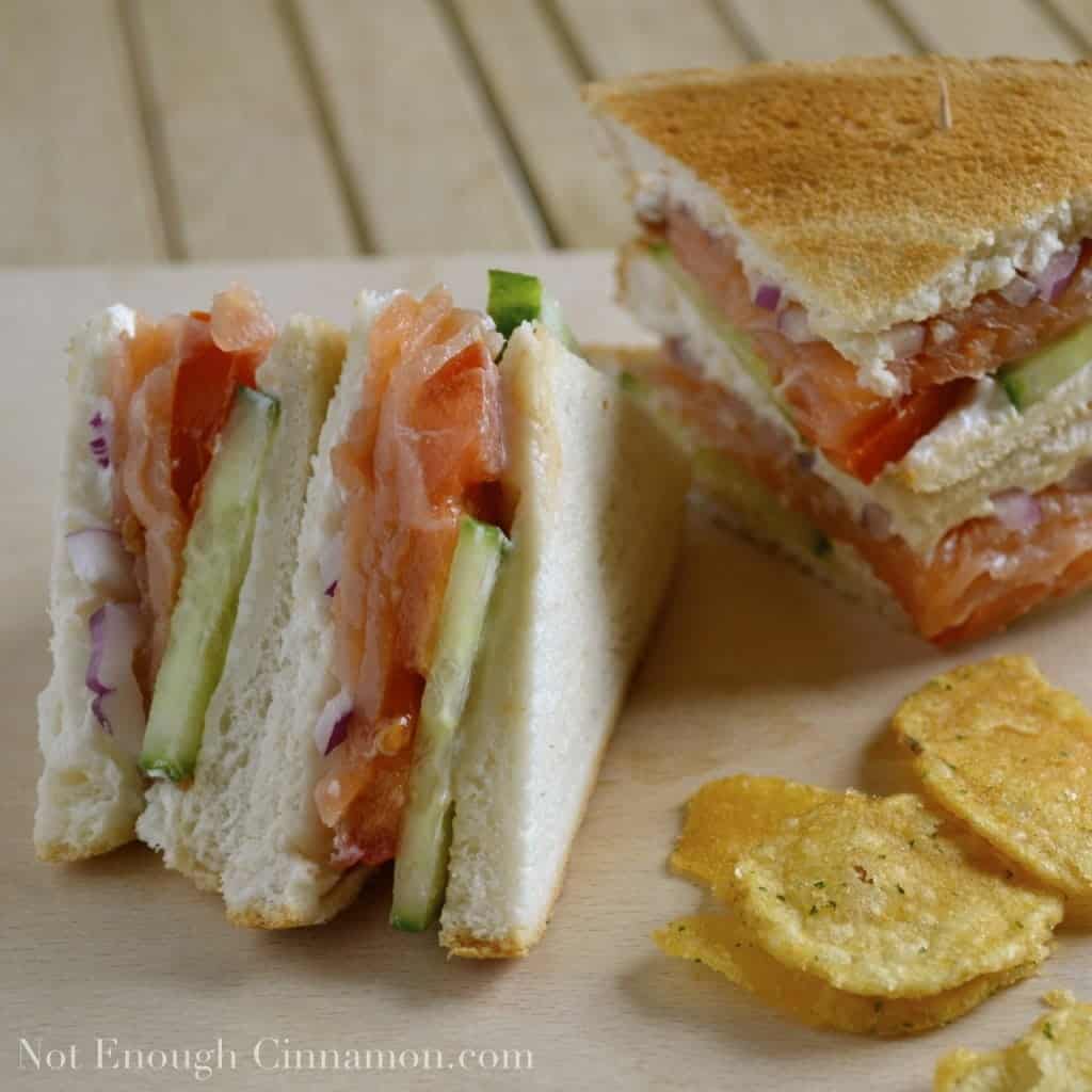 stacked smoked salmon club sandwiches with cucumber and red onion slices, cut into triangles and served with some potato chips on the side