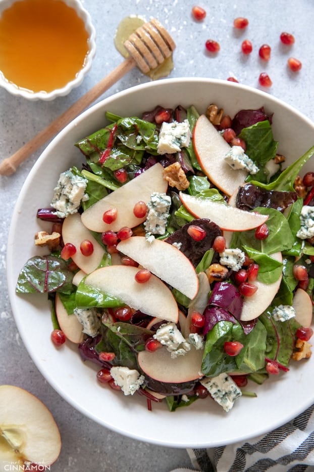 Apple and blue cheese salad in a white plate with pomegranate arils and honey on the side