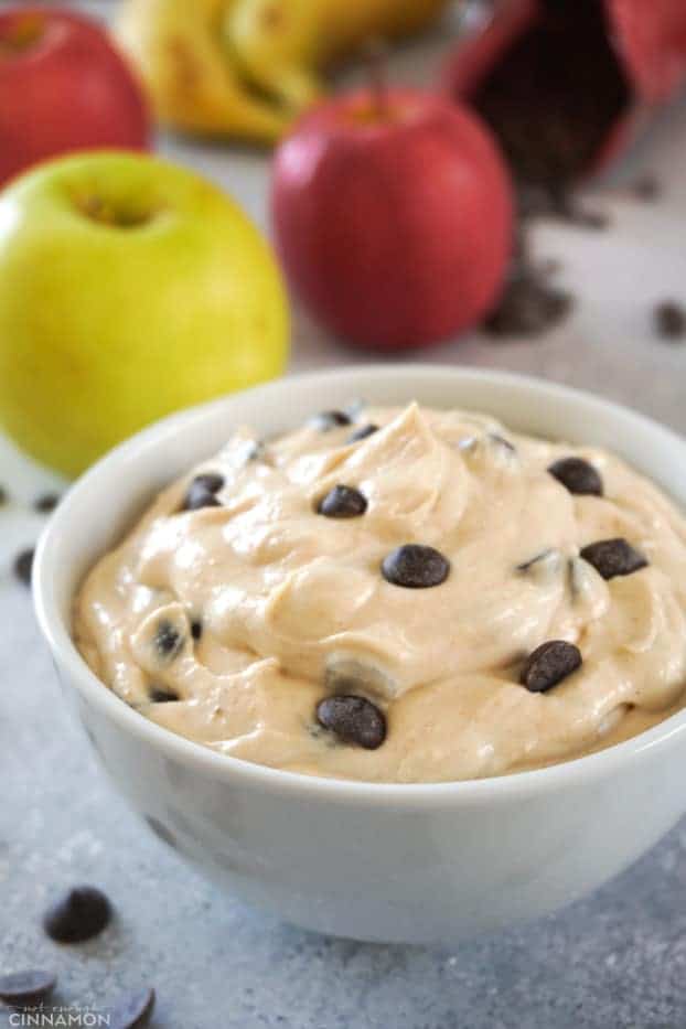 A white bowl of fruit dip with peanut butter and chocolate chips, with apples in the background.