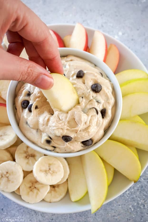 Apple dipped into peanut butter chocolate chips fruit dip in a white bowl, over a plate of fruit.