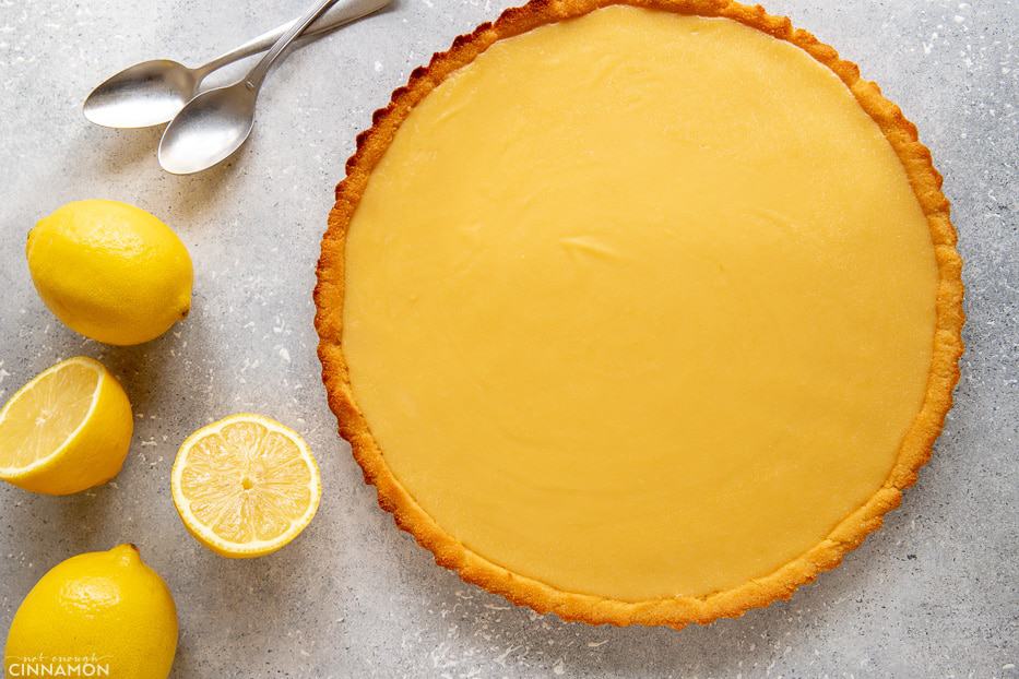 Whole paleo lemon curd tart with lemons and silver spoons on the side.