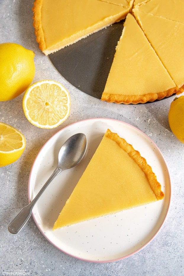 A slice of paleo lemon tart in a plate with a silver spoon and the whole tart sliced