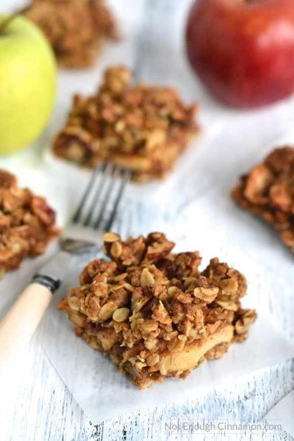 side view of healthy Apple Pie Oatmeal Bars arranged on a rustic white table with a fork and red and green apples in the background