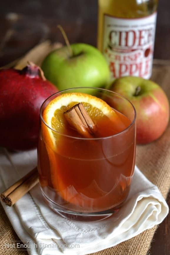 side view of a glass of pomegranate spiced apple cider with with an orange slice and cinnamon stick in it and apples in the background