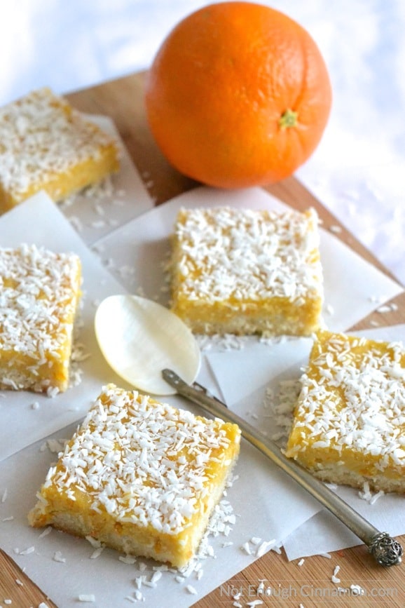Paleo Orange Curd Coconut Bars arranged on squares of white parchment paper with an orange in the background