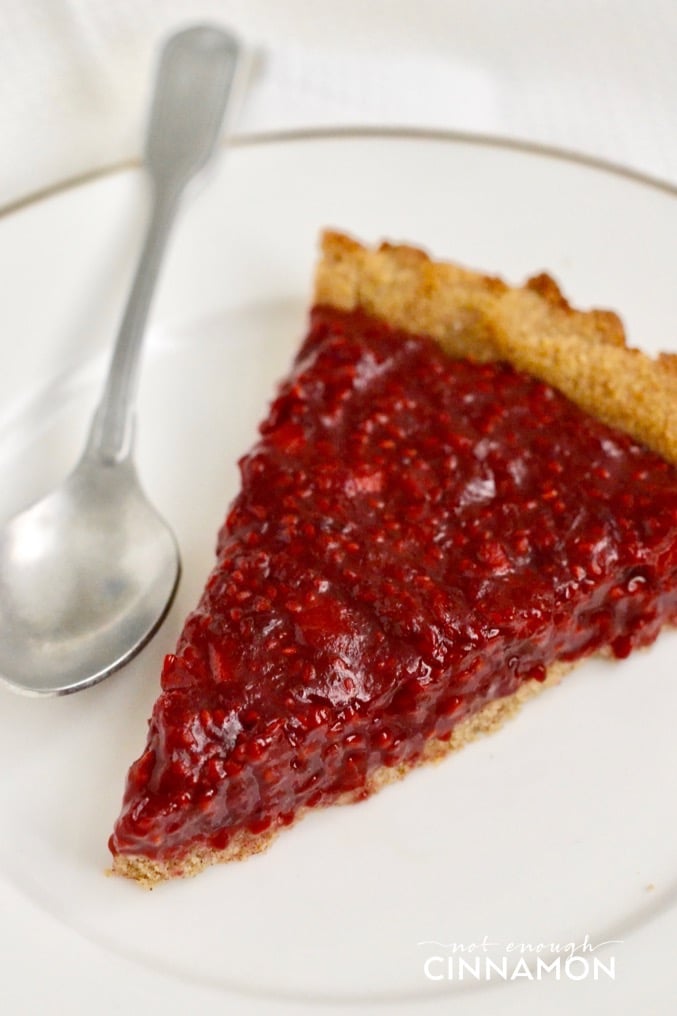 close-up of a slice of paleo raspberry cinnamon tart on a white plate with a spoon on the side
