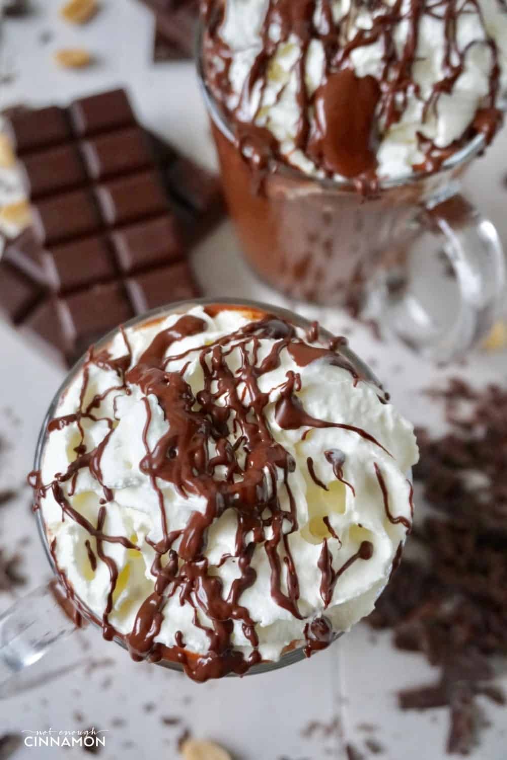 overhead shot of two tall glass mugs of peanut butter hot chocolate topped with whipped cream and chocolate sauce