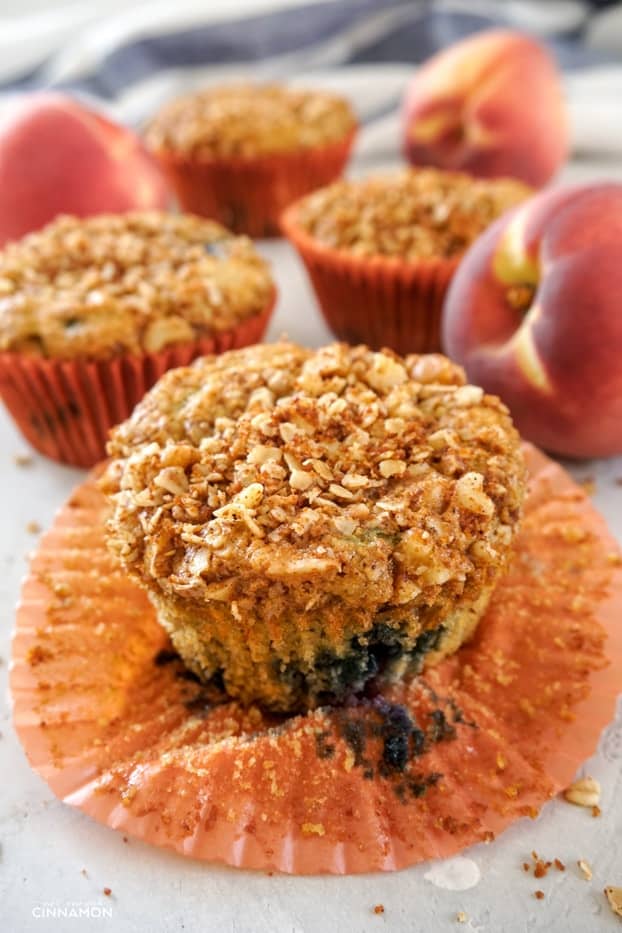 A peach and blueberry muffin with an orange liner opened up, and other muffins and peaches in the background. 