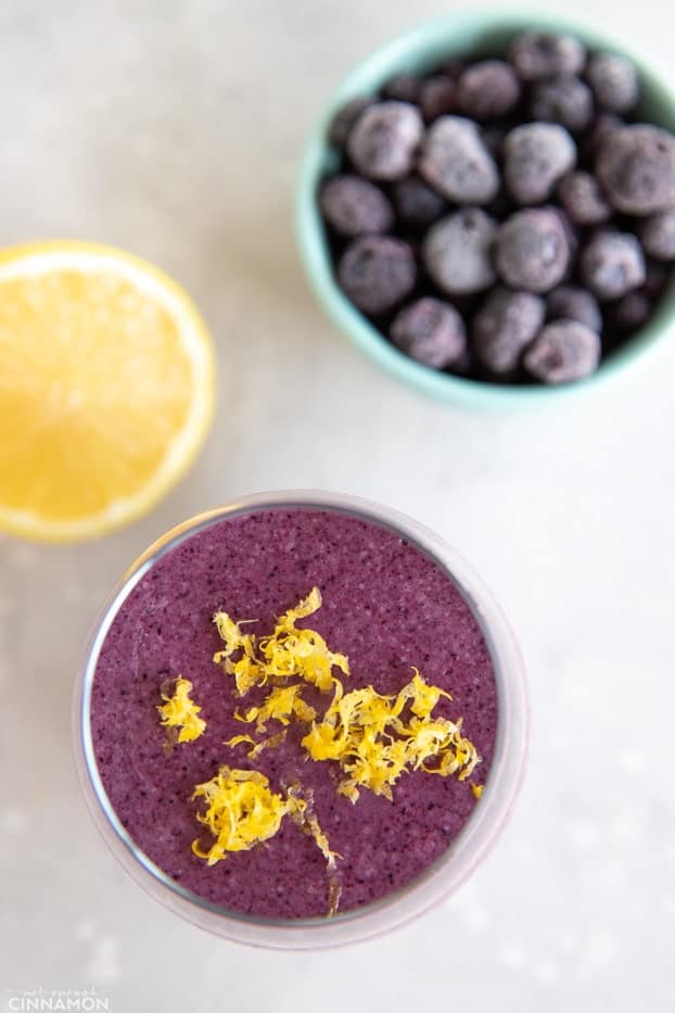 Lemon blueberry smoothie topped with lemon zest, with frozen blueberries and half a lemon on the side