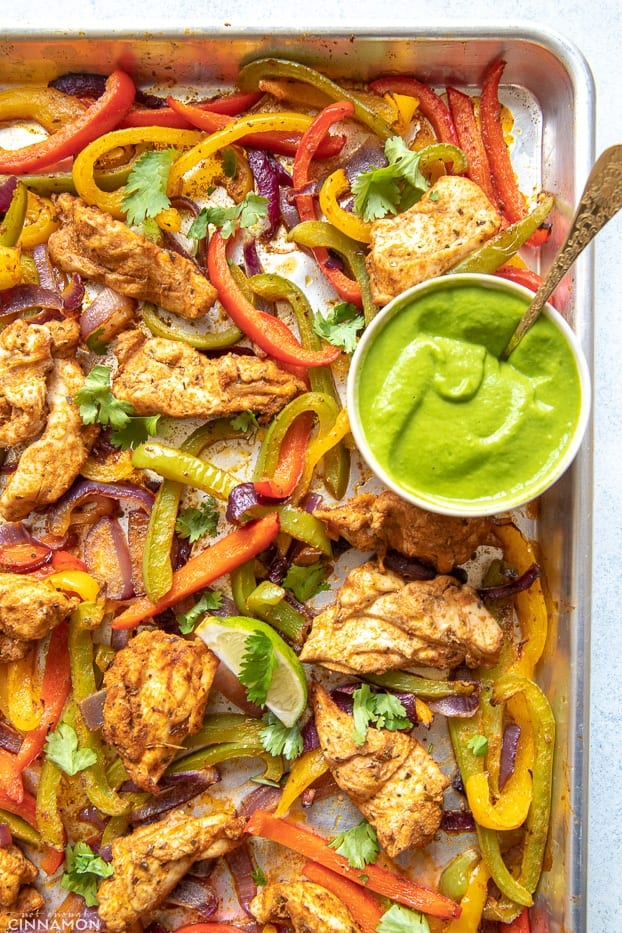 overhead shot of a sheet pan with low carb healthy oven baked chicken fajitas with a side dish containing creamy avocado sauce