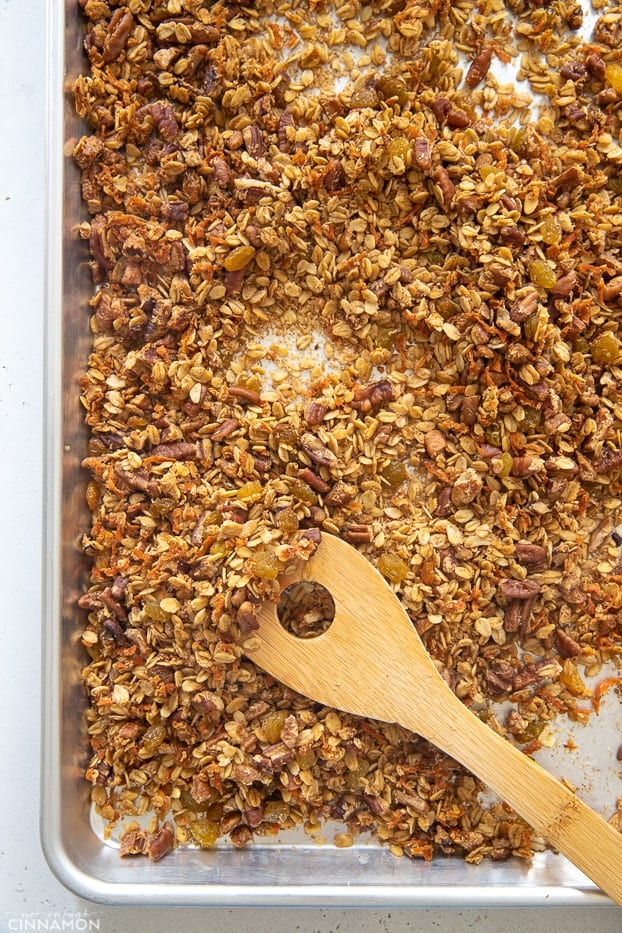 a wooden spoon stirring homemade gluten-free carrot cake granola on a sheet pan 