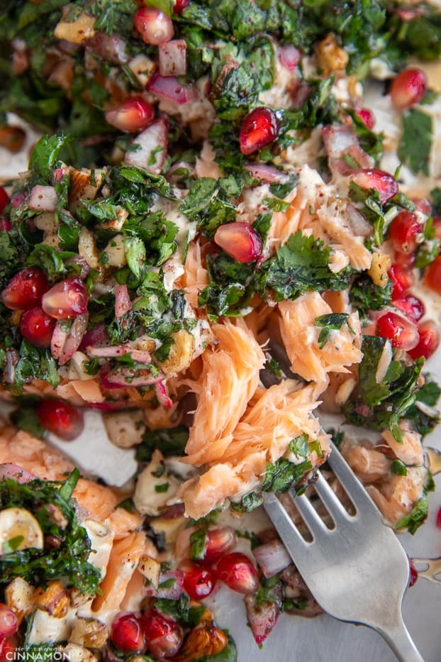 a fork taking a bite out of an oven baked salmon fillet topped with herbs and pomegranate seeds 