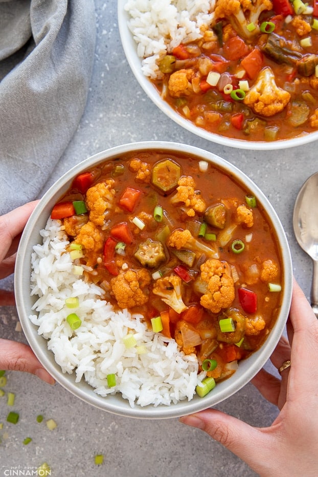 Zwei Hände halten eine Schüssel mit veganem Gumbo, serviert mit Reis und bestreut mit Frühlingszwiebeln