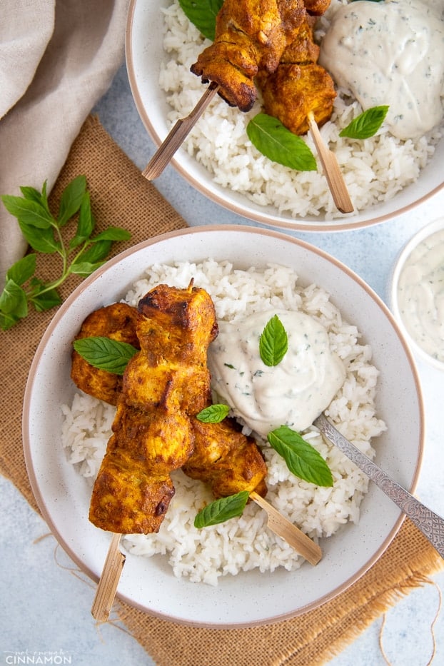 overhead shot of two bowls filled with rice and topped with baked tandoori chicken skewers served with dairyfree yogurt dip