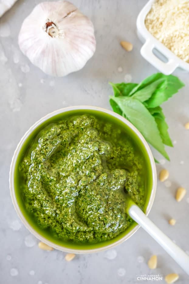 Bright green basil pesto in a small bowl with a white spoon, some basil leaves, a few pine nuts, a garlic clove and grated parmesan cheese.