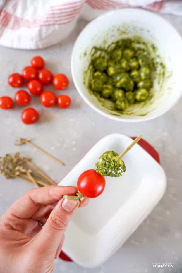 Hand holding a tomato, pesto and mozzarella skewer being made, with cherry tomatoes, pesto covered mozzarella balls and wooden skewers in the background.