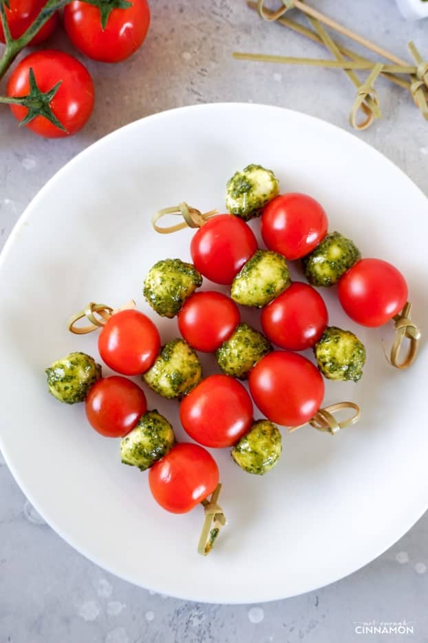 Five tomato, pesto and mozzarella skewers on a white plate, with grape tomatoes and wooden skewers in the background. 