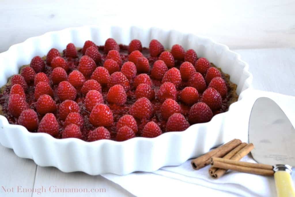 Skinny Raspberry Tart with raspberry jam and fresh raspberries on whole wheat cinnamon crust in a white tart dish with some cinnamon sticks on the side