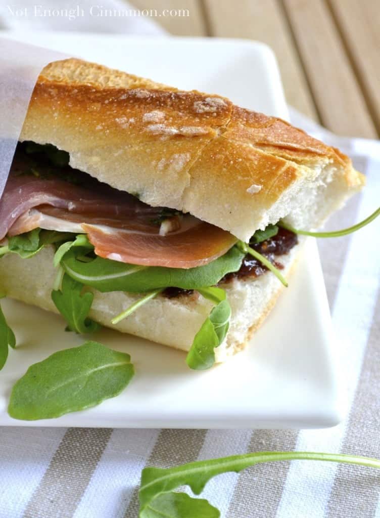 side view of a French baguette prosciutto sandwich with pesto, arugula and fig chutney halfway wrapped in sandwich paper and served on a white plate