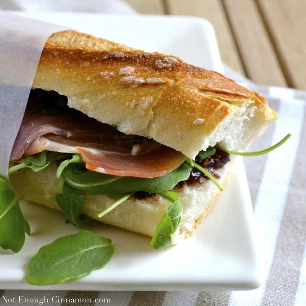 side view of a prosciutto sandwich with pesto, arugula and fig chutney served on a white plate with a strip of sandwich paper holding it together