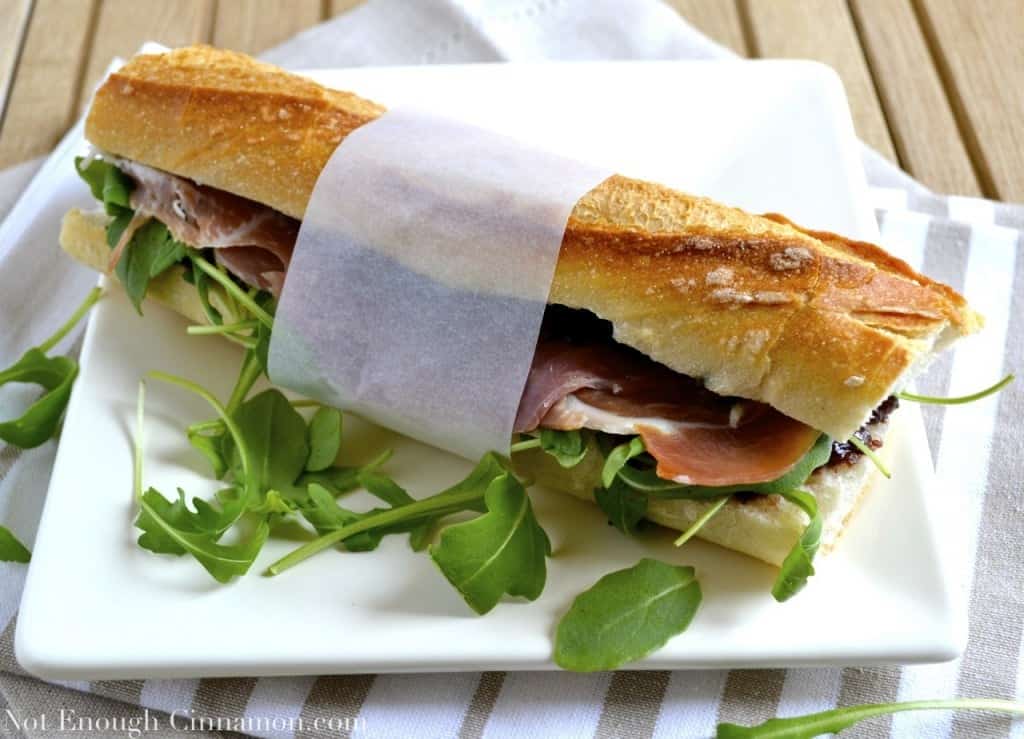 French baguette prosciutto sandwich with pesto, arugula and fig chutney with a strip of white sandwich paper around the middle, served on a white plate