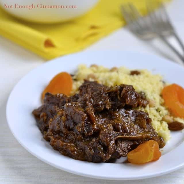 Delicious Lamb Tagine with dried Apricots served with a side of raisin studded couscous on a white plate with a bowl of couscous in the background