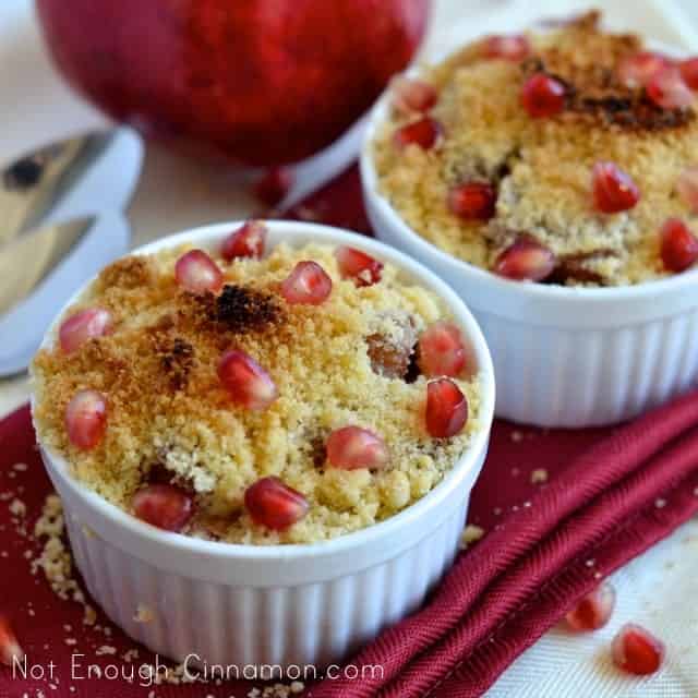 Individual Pear and Pomegranate Crumbles in white ramekins sprinkled with pomegranate seeds on a red festive napkin with a pomegranate in the background