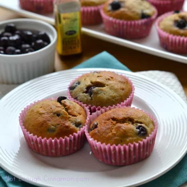 Three Blueberry Banana Bread Muffins on a white plate with a tray of muffins and fresh blueberries in the background