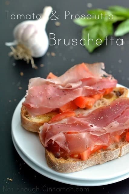 2 pieces of Prosciutto Tomato Bruschetta on a white plate on a black tabletop with garlic and basil in the background