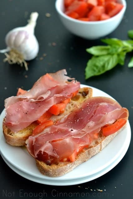 2 pieces of Prosciutto Tomato Bruschetta on a white plate placed on a black tabletop with garlic and basil in the background