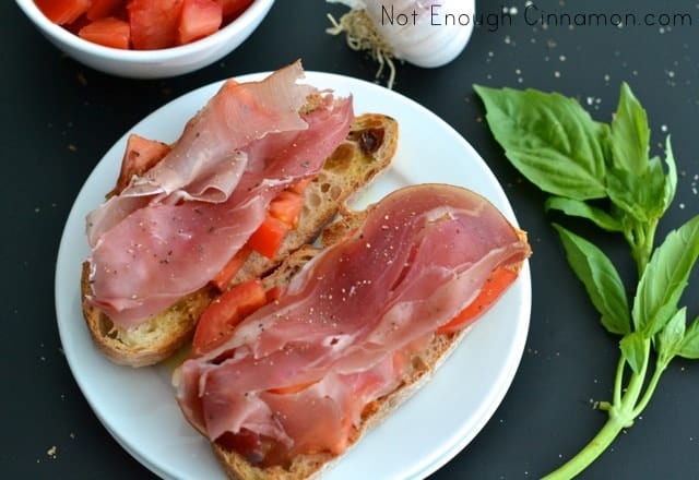 2 pieces of Prosciutto Tomato Bruschetta on a white plate on a black tabletop with garlic and basil in the background