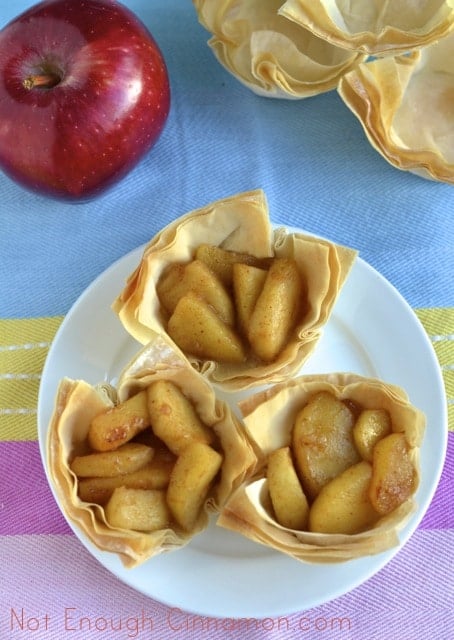 three Caramelized Apple Phyllo Cups on a white plate with an apple and empty baked phyllo cups in the background