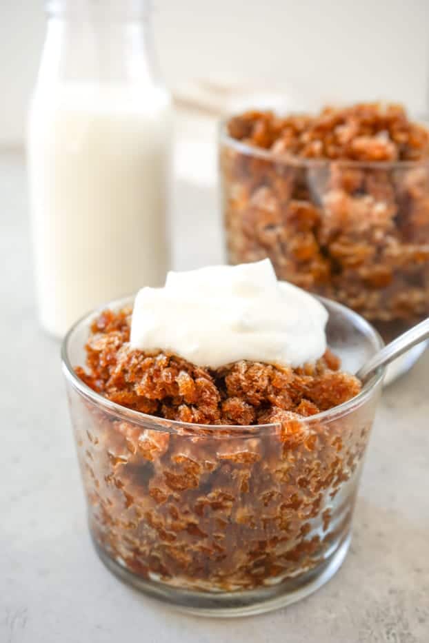 Coffee granita in a transparent glass, with a metal spoon and topped with whipped cream. Another glass of granita and a small bottle of milk in the background. 