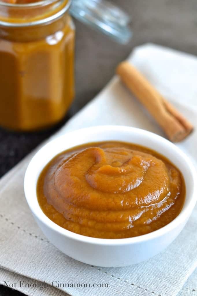  Slow Cooker Applesauce with pumpkin and pumpkin spices served in a small white bowl with a cinnamon stick and a jar of slow cooker applesauce in the background