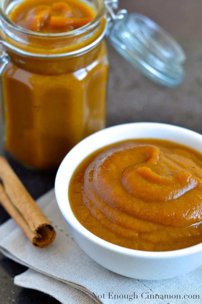  Slow Cooker Applesauce with pumpkin and pumpkin spices served in a small white bowl with a cinnamon stick and a jar of slow cooker applesauce in the background