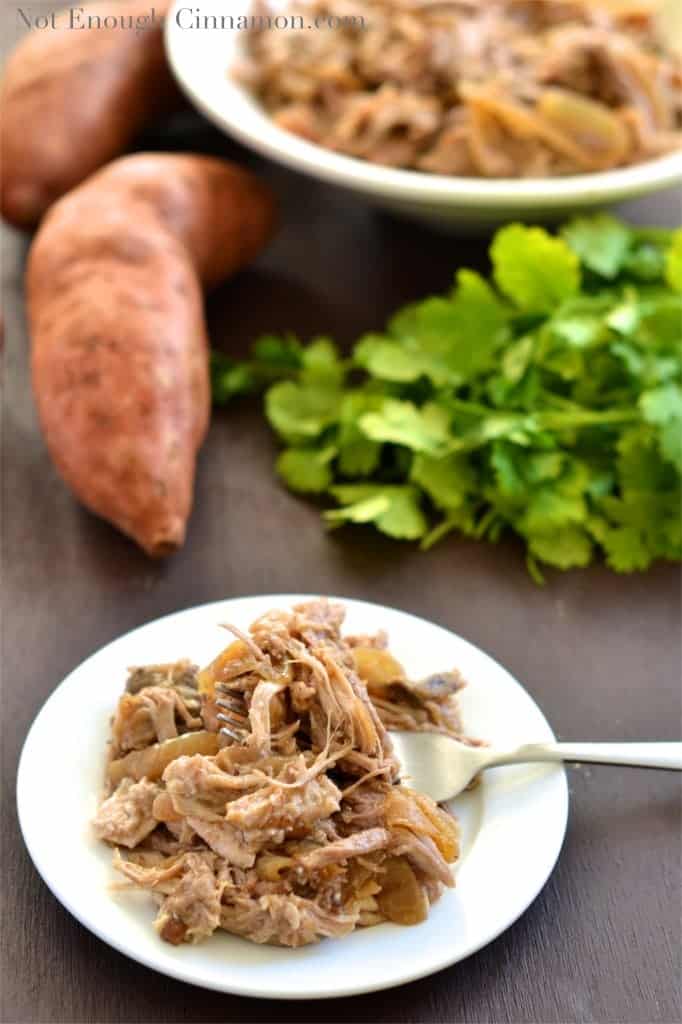 a serving of Slow Cooker Pulled Pork with Apples and Onions served on a small white plate with sweet potatoes and cilantro in the background