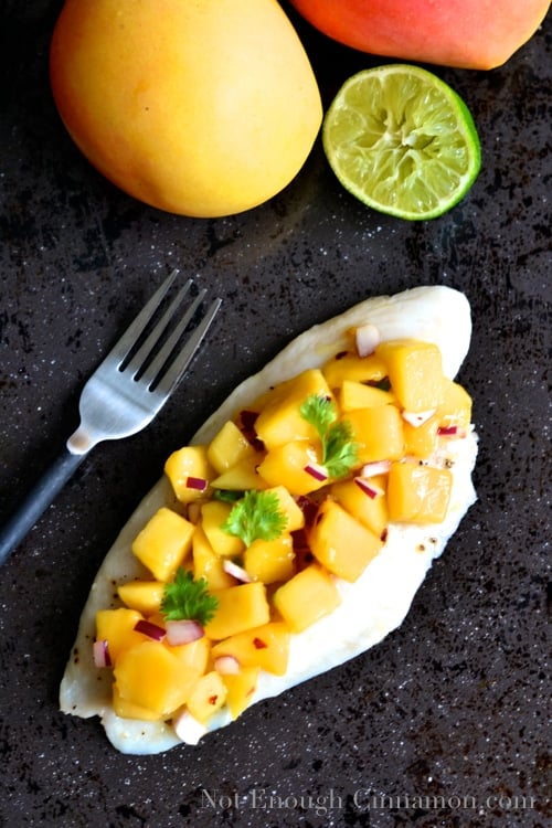 overhead shot of white fish fillet topped with mango lime salsa next to a fork, with some mangoes and half a lime in the background