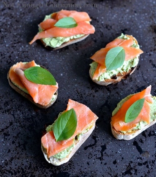 Smoked Trout and Basil Pesto Cream Cheese Crostini topped with fresh basil leaves and arranged on a black surface