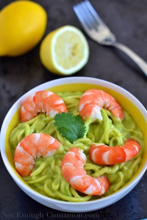Zucchini noodles with creamy avocado sauce and shrimps served in a white bowl with lemons and a fork in the background