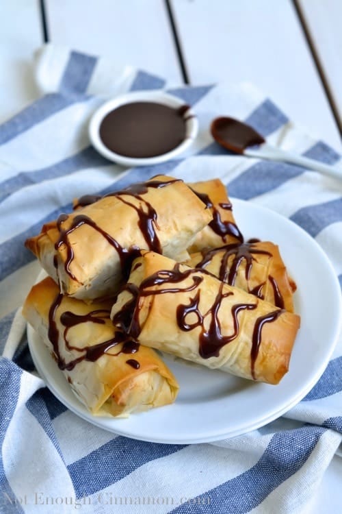 Baked Chocolate and Banana Spring Rolls drizzled with melted chocolate and served on a white plate with a small dish of melted chocolate in the background