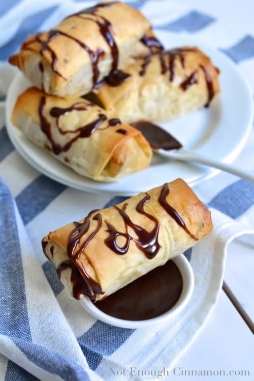 a Baked Chocolate and Banana Spring Roll resting on a small dish with melted chocolate with more sweet spring rolls in the background