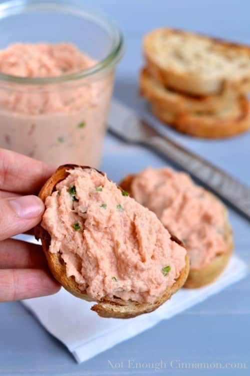 a hand holding a slice of toasted baguette topped with a generous amount of French Salmon Rillettes made from smoked and fresh salmon
