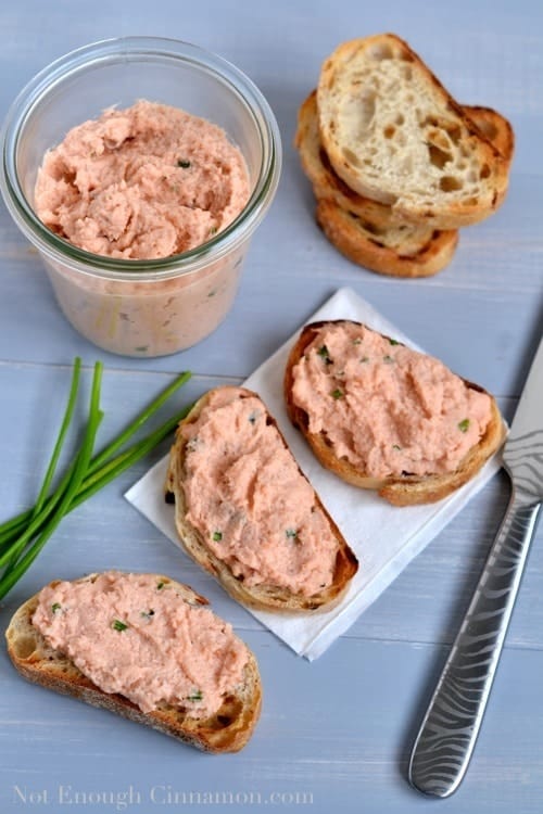 overhead shot of three slices of toasted baguette topped with homemade salmon rillettes with some chives, baguette slices and a half-filled jar or rillettes on the side