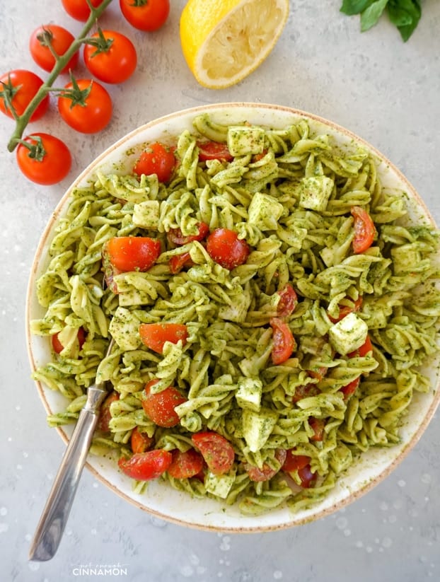 Fusilli pasta, cherry tomatoes, mozzarella with a pesto dressing in a large white bowl, with a large spoon, tomatoes and half a lemon in the background.