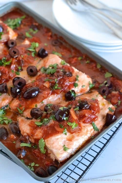 Oven Baked Salmon in a rich tomato olive Provencal Sauce served in a casserole dish with some plates in the background