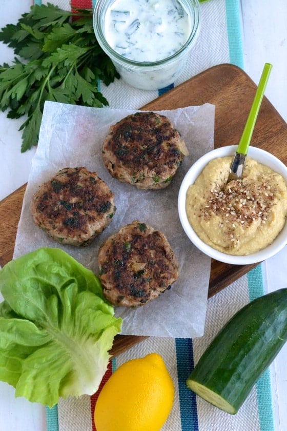 overhead shot of Middle Eastern Lamb Burger patties arranged on a wooden board with a small bowl if hummus and some homemade tzatziki on the side
