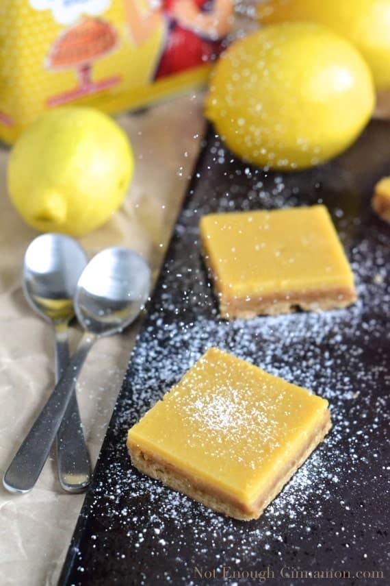 powdered sugar being sprinkled on top of easy lemon bars arranged on a black tray with lemons in the background