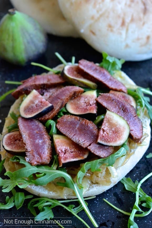 close-up of Lamb sandwich topped with arugula, caramelized onions and drizzled with balsamic reduction, served on a black tabletop