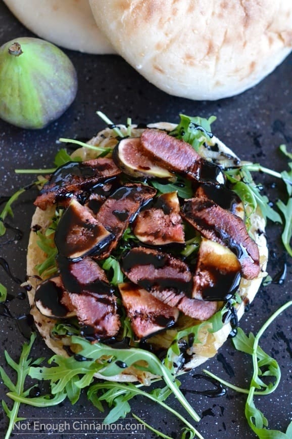 an open Lamb sandwich topped with sliced figs, arugula, caramelized onions and drizzled with balsamic reduction, served on a black tabletop