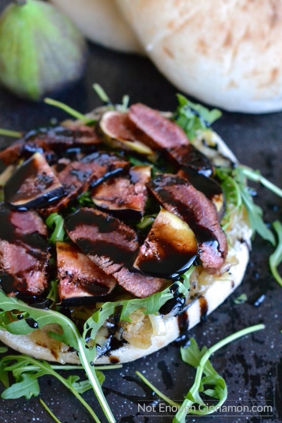 an open Lamb sandwich topped medium rare lamb slices, figs, arugula, caramelized onions and drizzled with balsamic reduction, served on a black tabletop